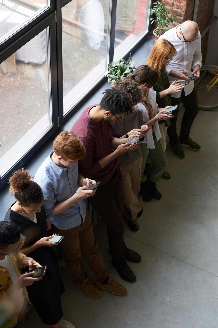 A group of diverse office workers standing by a window using smartphones, capturing modern workplace connectivity.. fauxels: https://www.pexels.com/@fauxels
