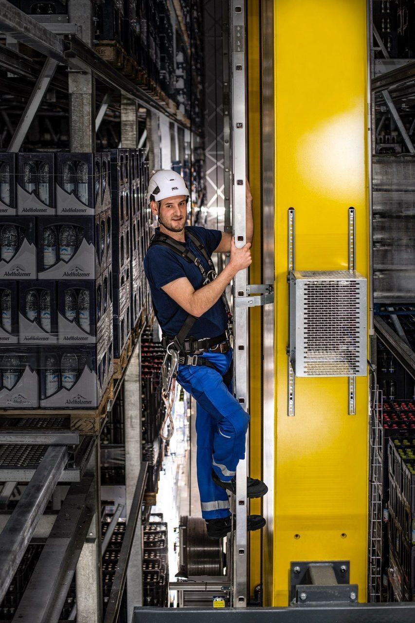Man in safety gear climbing a ladder in a high-bay warehouse, viewed vertically.