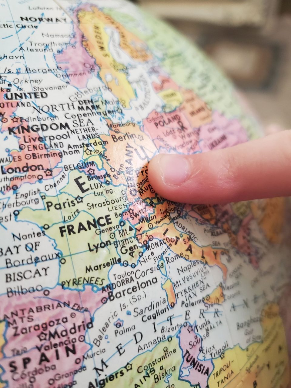 A child&#x27;s finger points to Germany on a globe. The bright colors of the map has bold black letters indicating places in the world.
