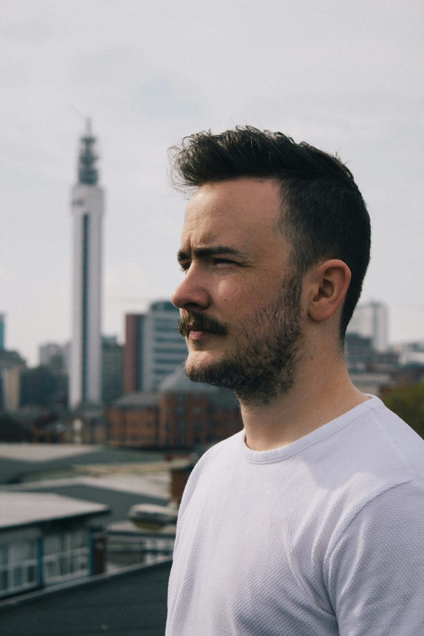 Profile of man with beard and mustache, white shirt, city skyline and tall tower.