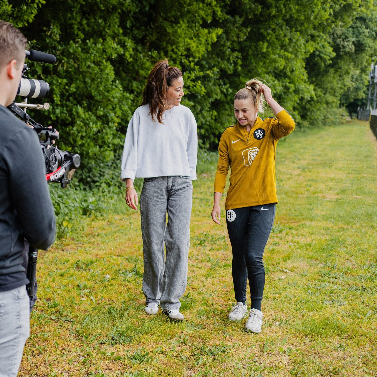 Two women walk and chat on grass, one in a yellow sports top, filmed by a cameraperson.