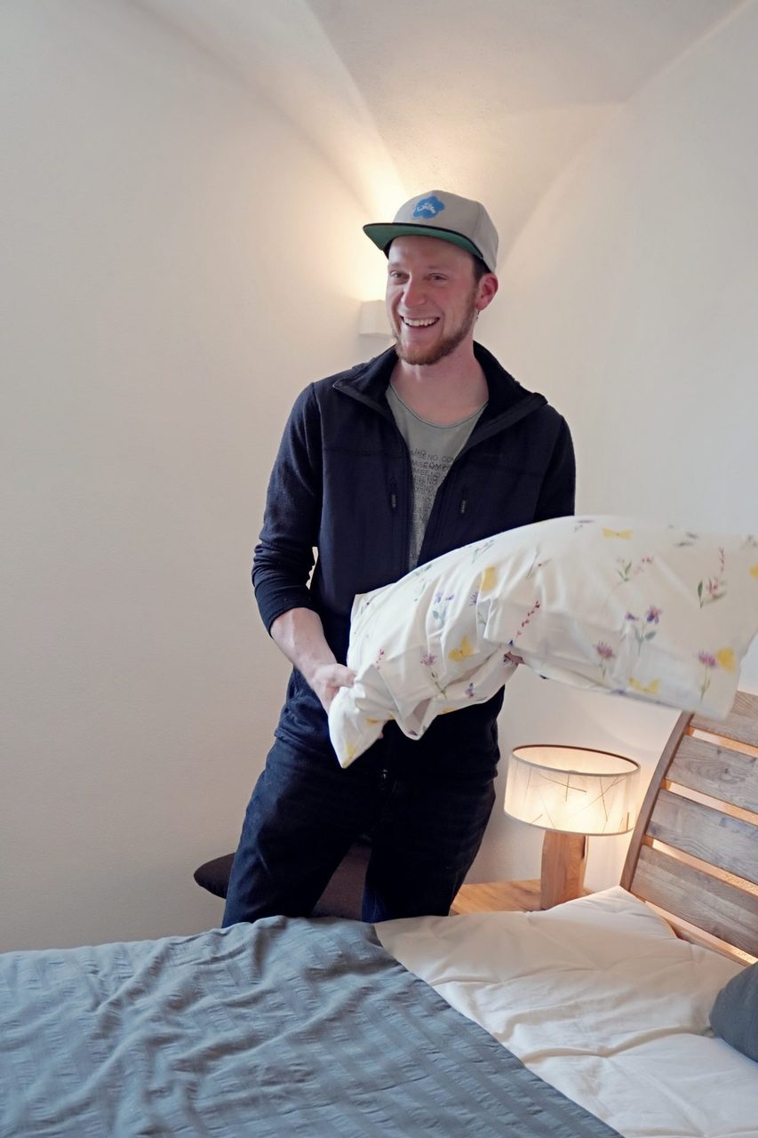 Smiling man in a cap holding a floral pillow in a bedroom.