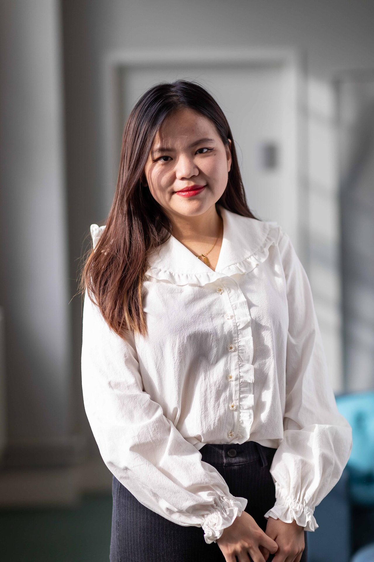 A smiling Asian woman with long dark hair, wearing a white ruffled shirt and red lipstick.