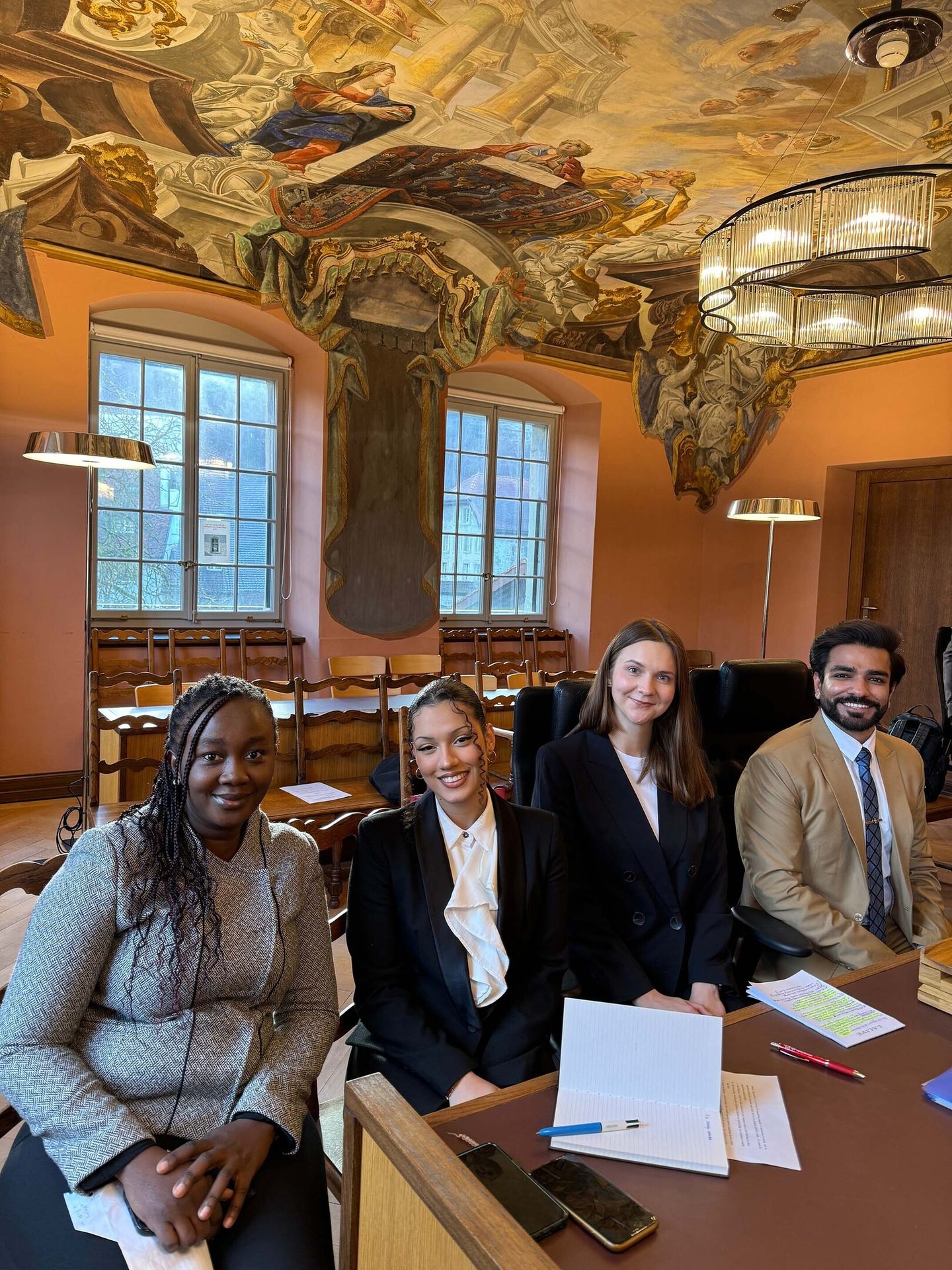 Four diverse people in formal attire at a table in a room with an ornate, painted ceiling.