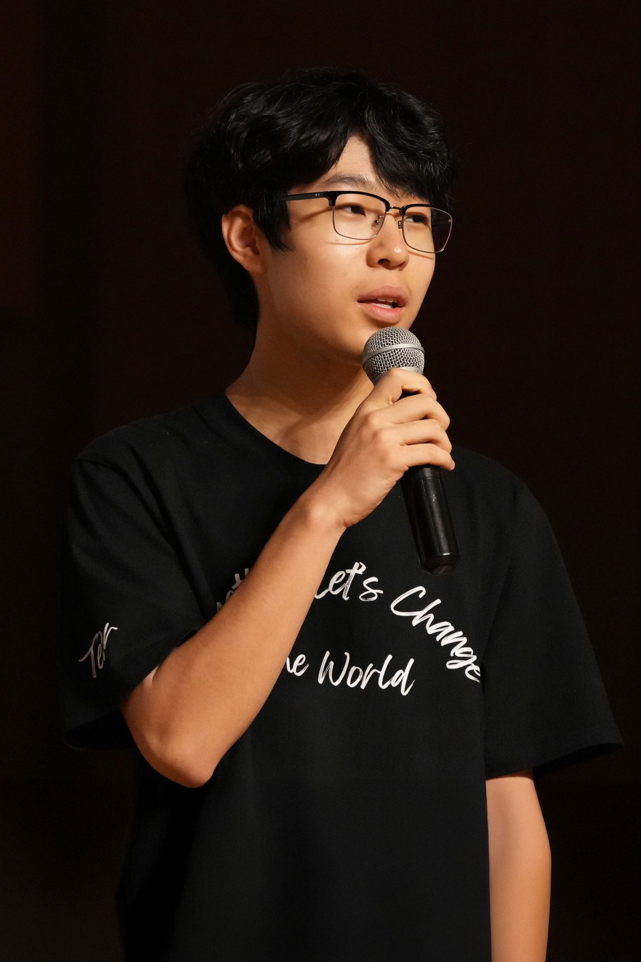 A young person with glasses speaks into a microphone, wearing a black "Let's Change the World" t-shirt.