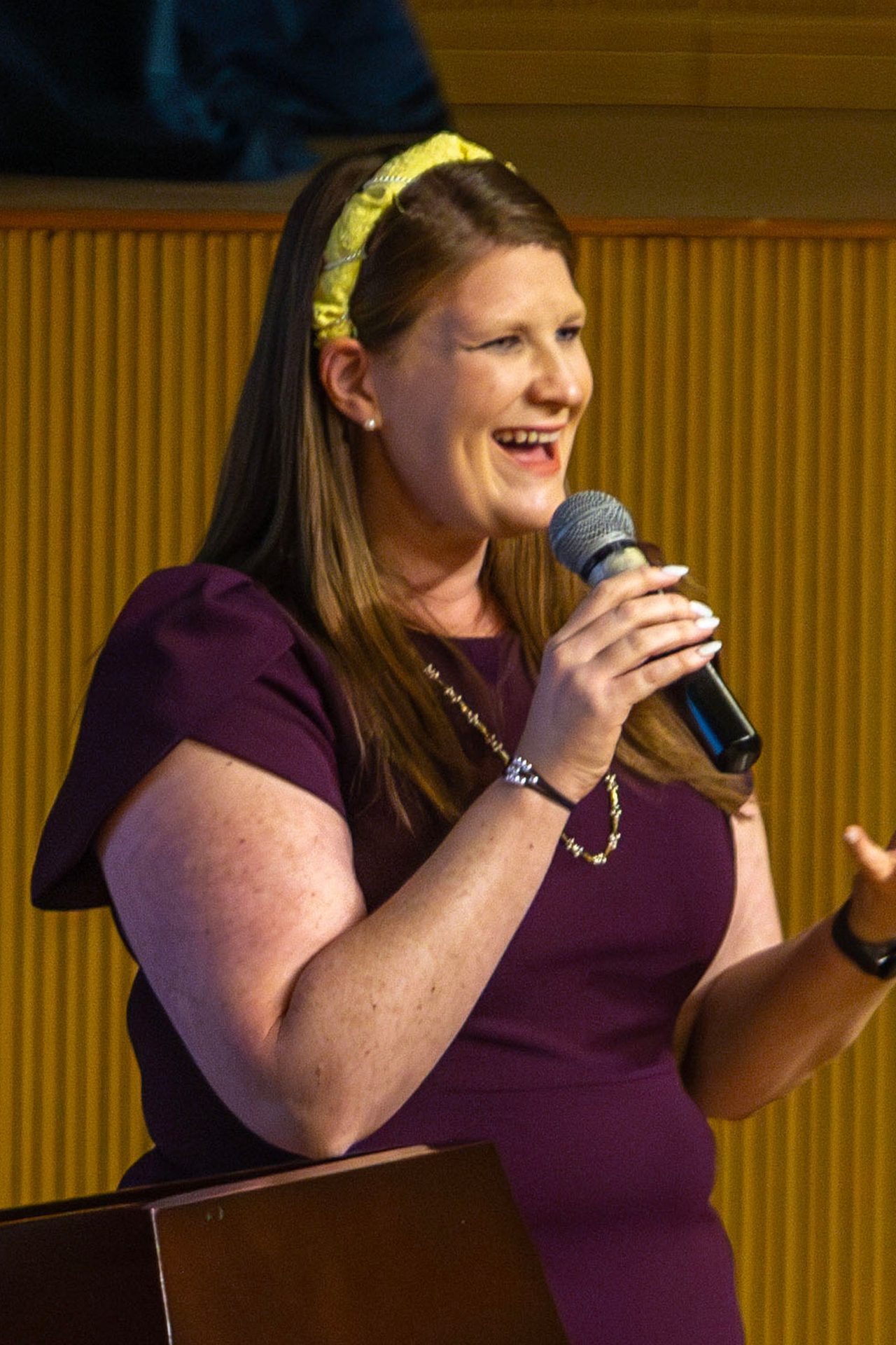 A smiling woman in a purple dress and yellow headband speaks into a microphone.