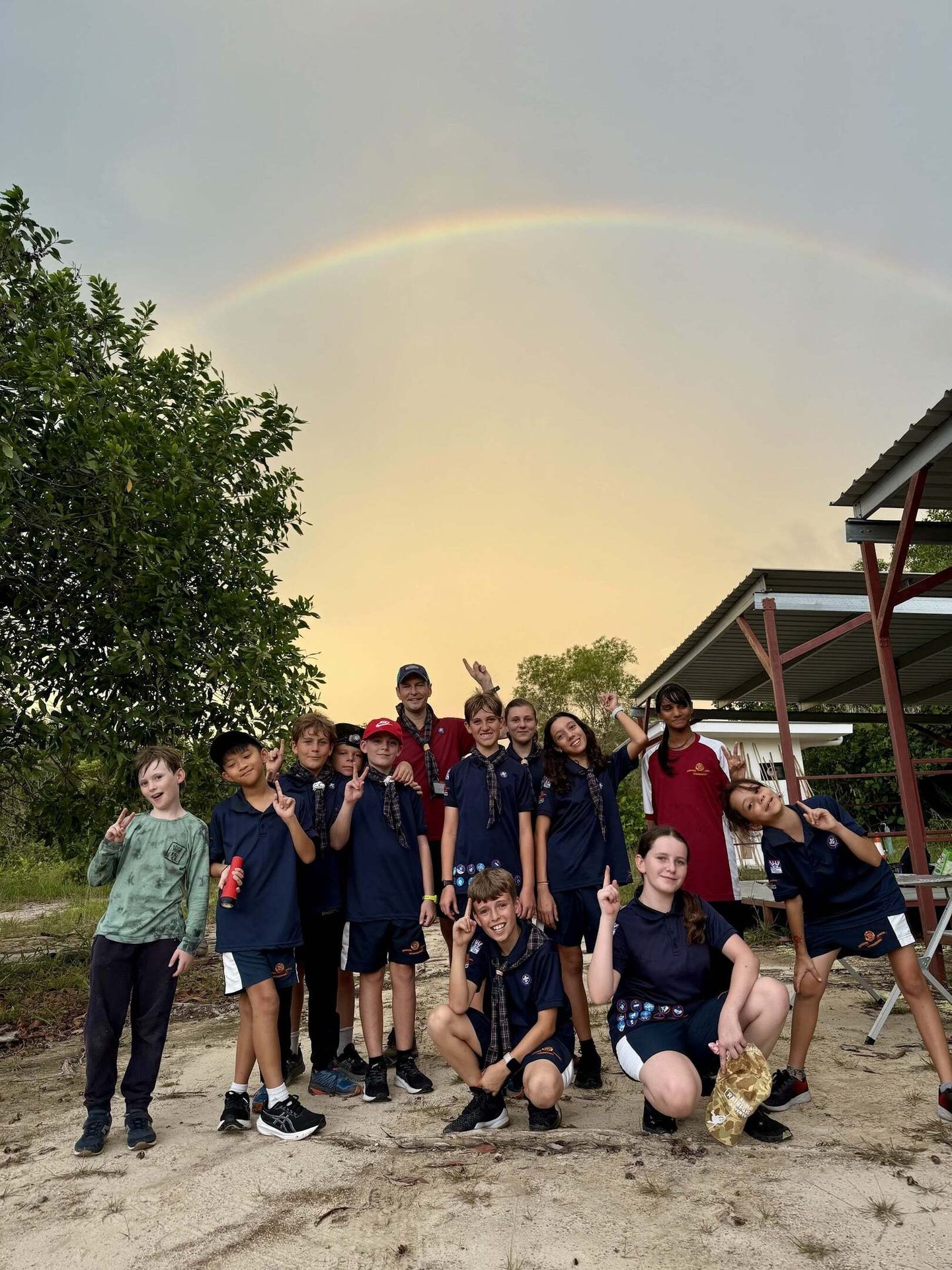 A group of children and adults pose outdoors under a large rainbow in the sky.