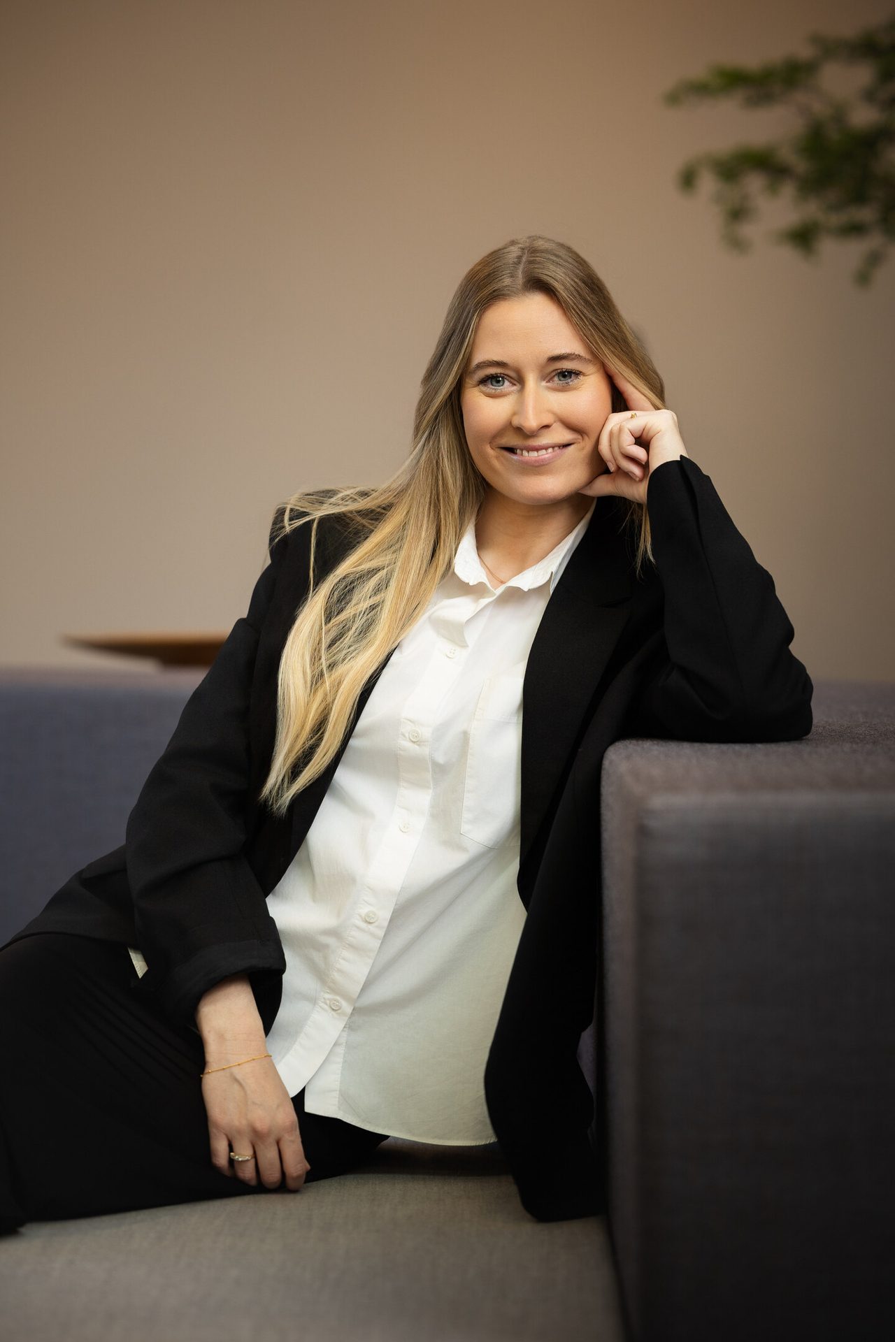 Smiling blonde woman in black blazer & white shirt, seated on a couch.
