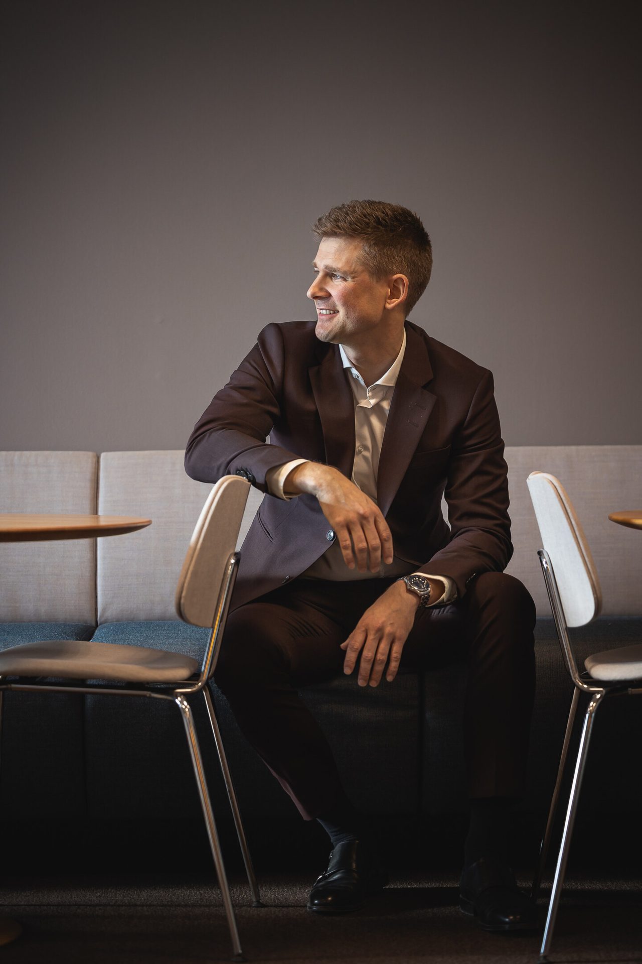 A smiling man in a dark suit sits casually on a sofa, looking to his right in a relaxed setting.