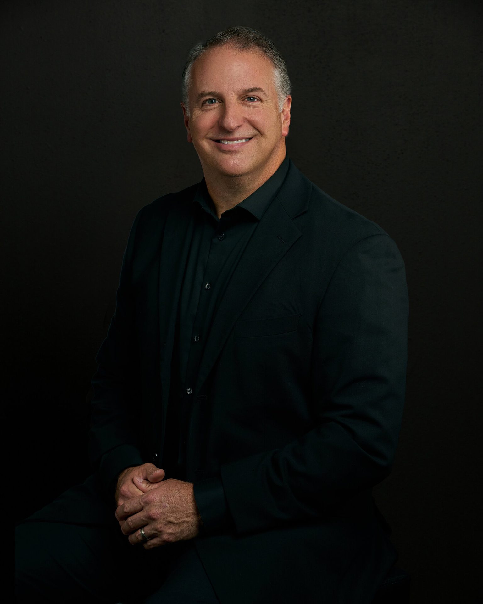 Smiling man in dark suit, hands clasped, dark background.