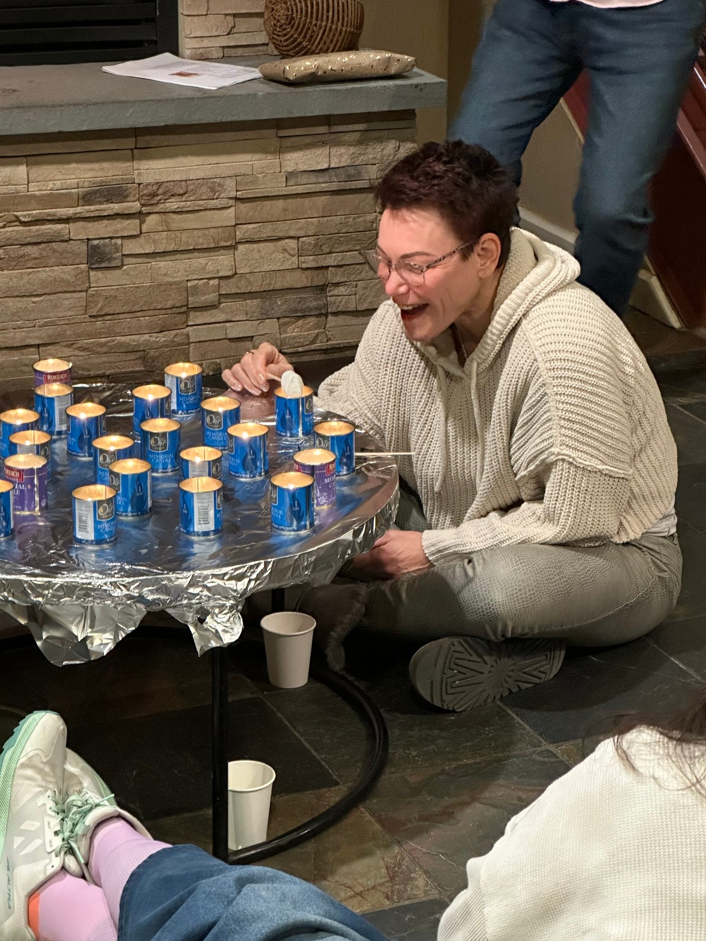 A smiling woman lights memorial candles on a foil-covered table in front of a stone fireplace.