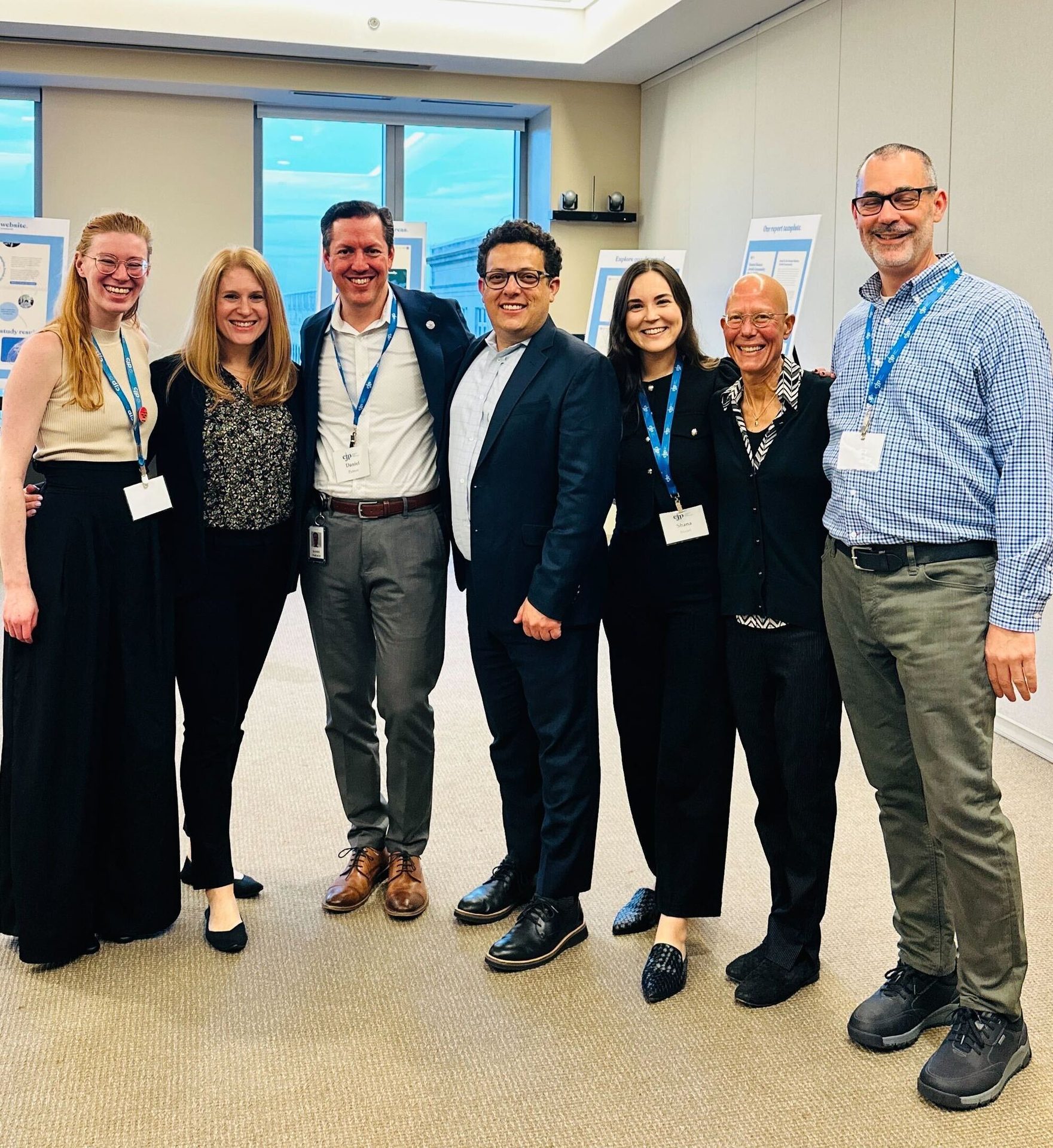 Group of seven smiling people indoors, wearing lanyards.
