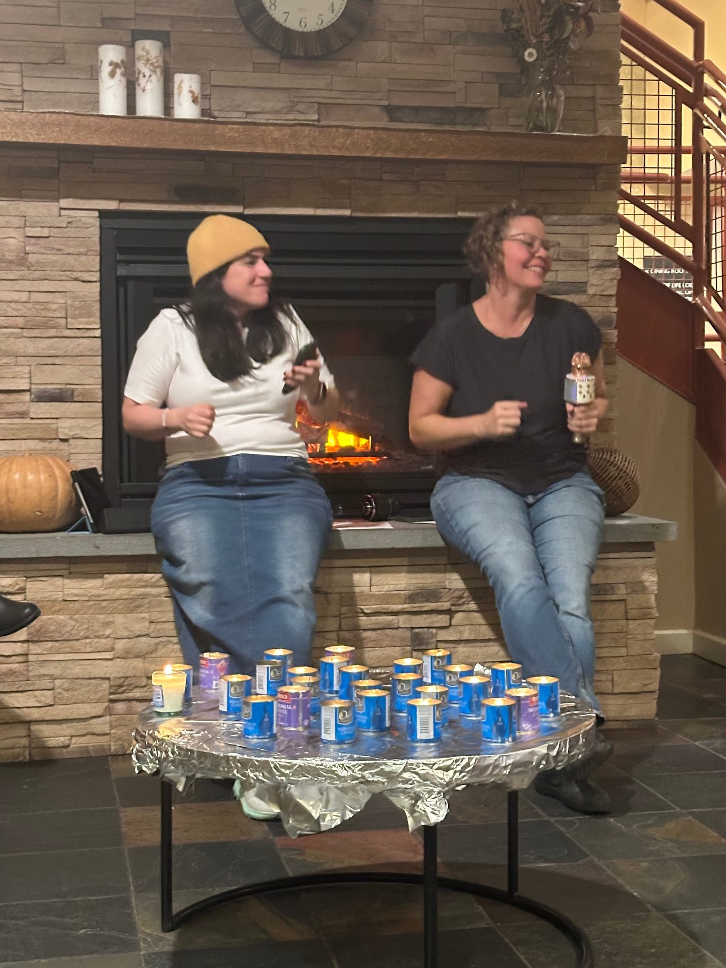 Two women sing by a fireplace, with lit candles on a table in front of them.