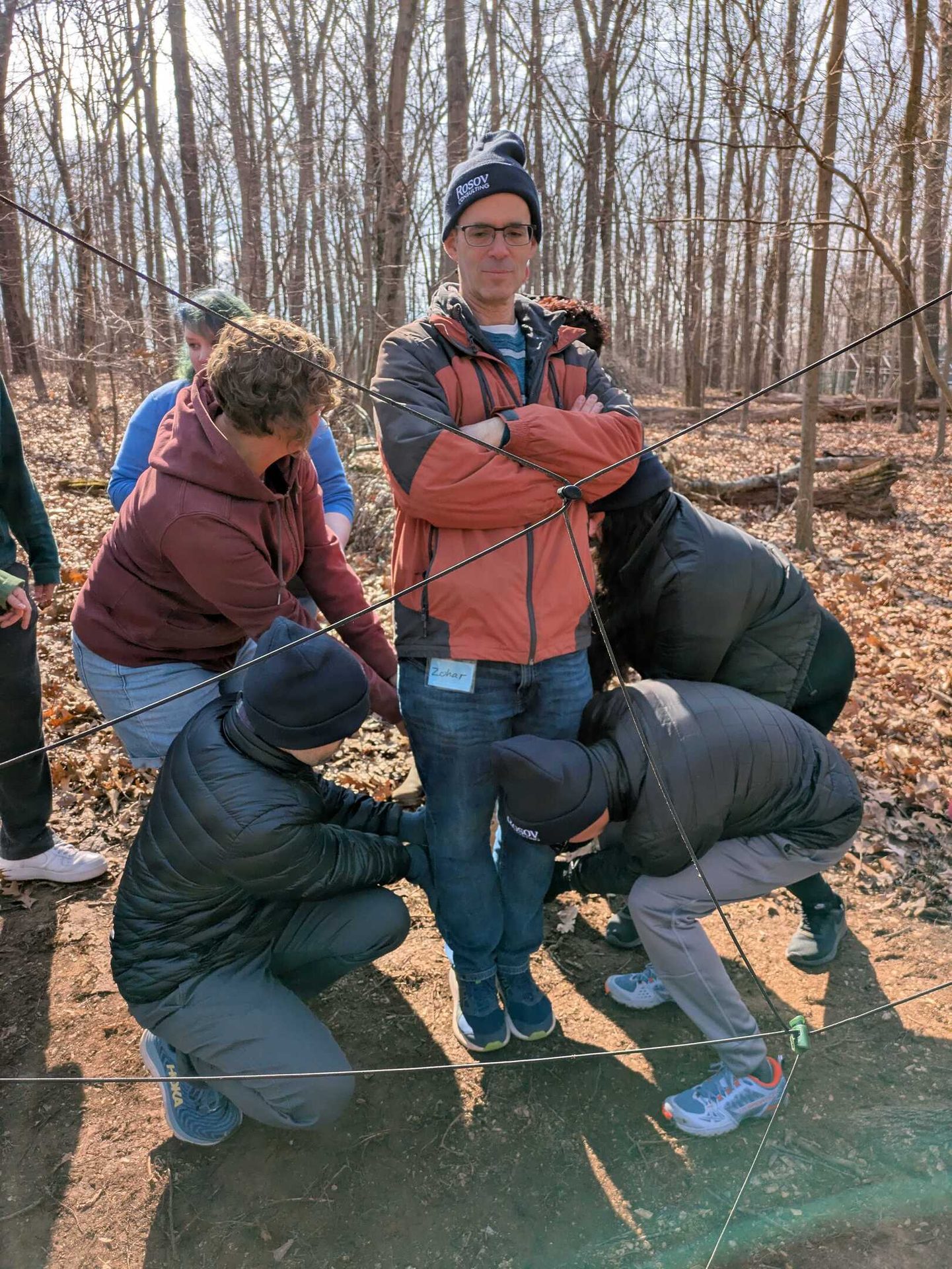 Group in a forest doing a rope activity, focused on a man in the center.