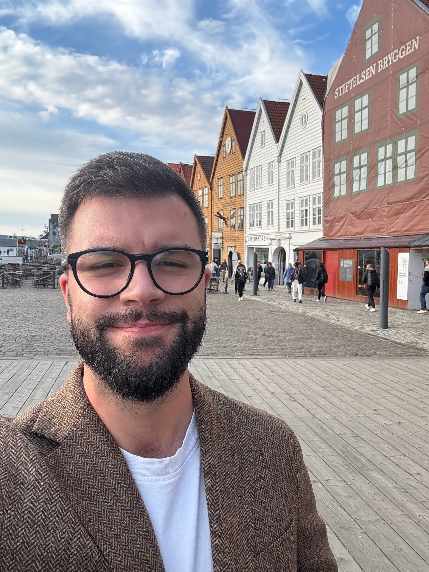 Smiling man with beard & glasses takes selfie at Bryggen, Bergen, with historic buildings.