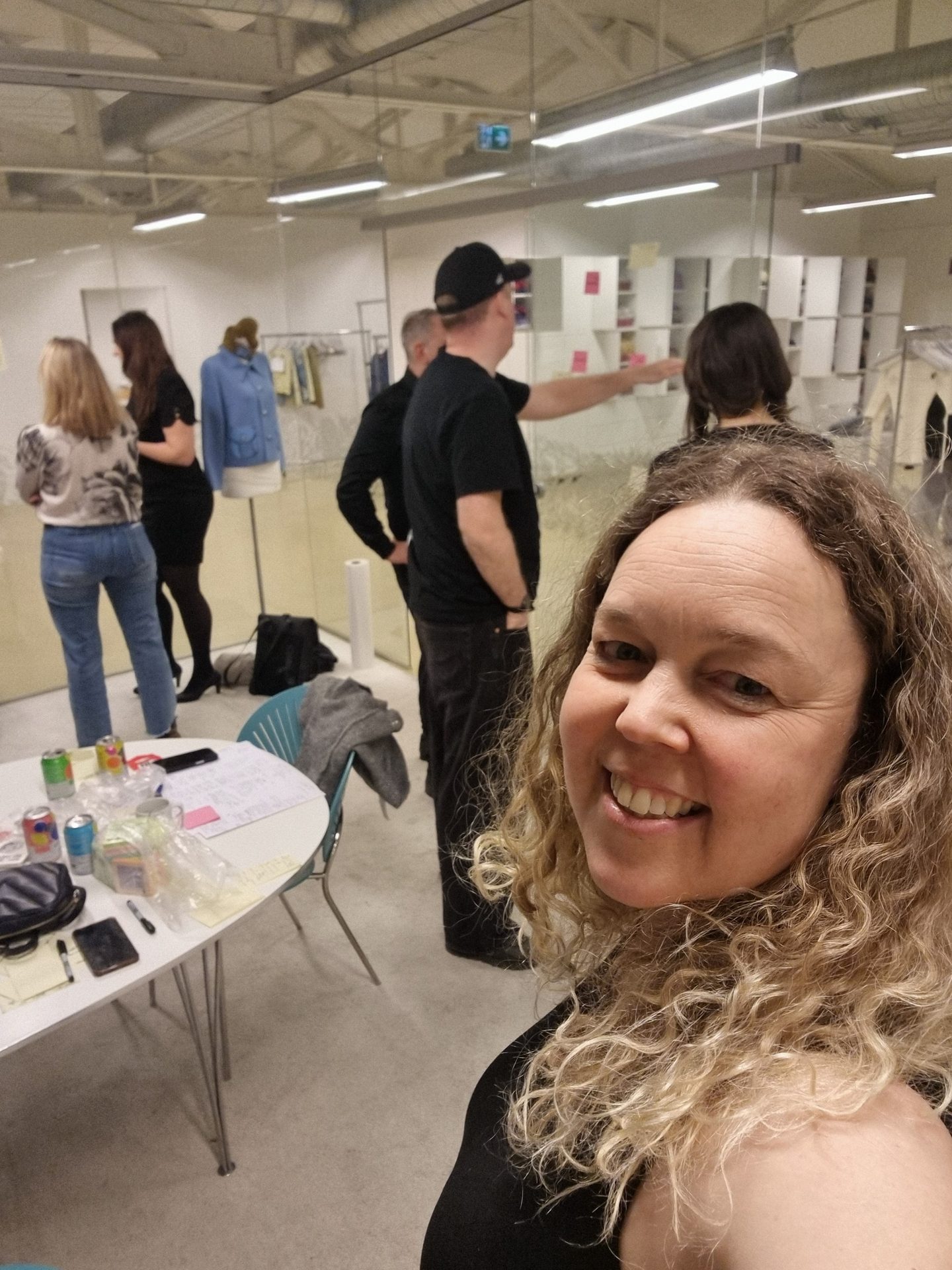A smiling woman with curly hair takes a selfie in a brightly lit room with a group of people in the background.