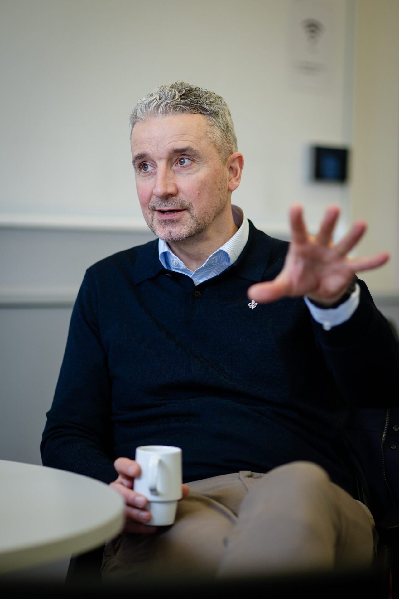 Man with gray hair, wearing a dark blue sweater, holding a cup and gesturing.
