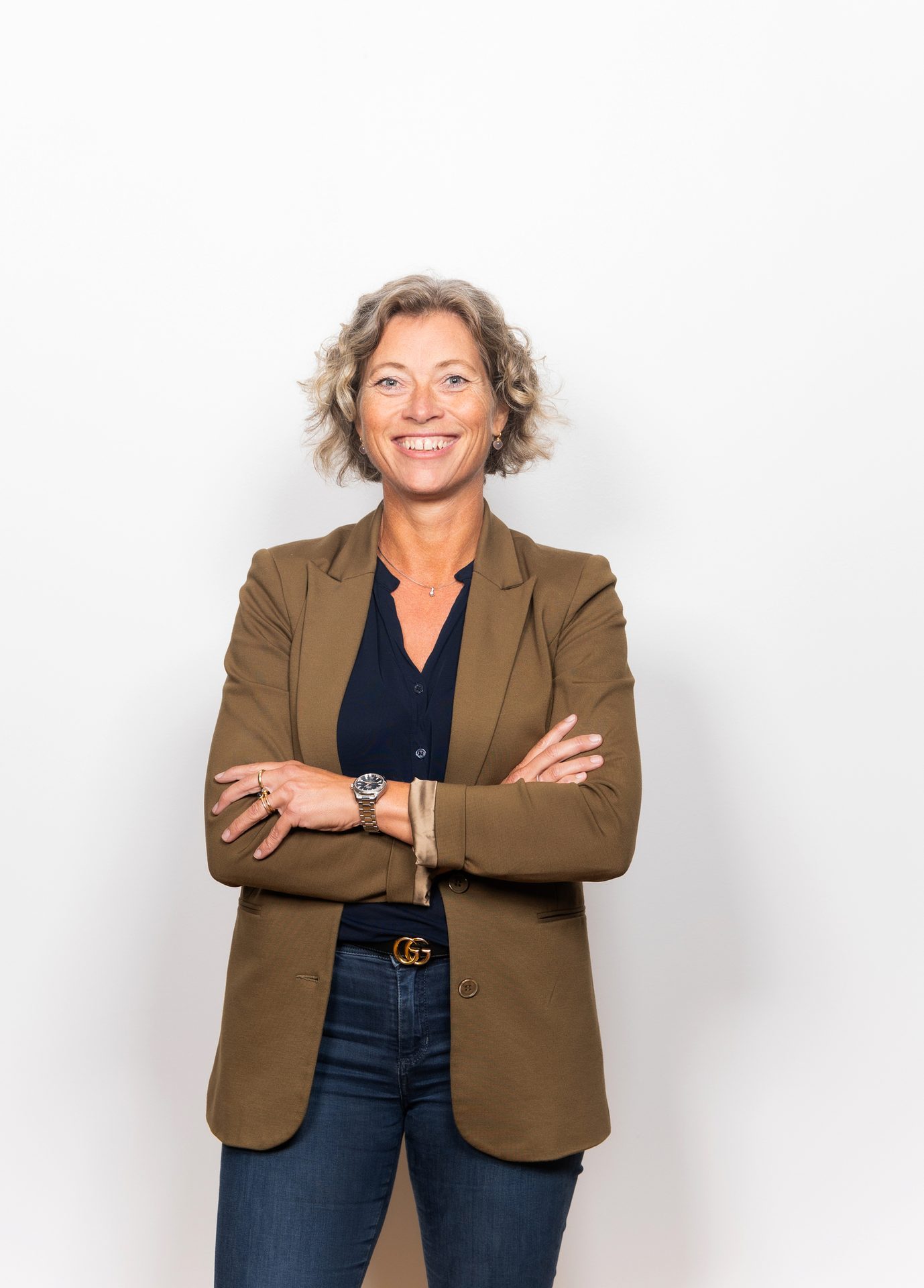 Smiling woman with curly hair, brown blazer, navy shirt, jeans, and a Gucci belt. Arms crossed.