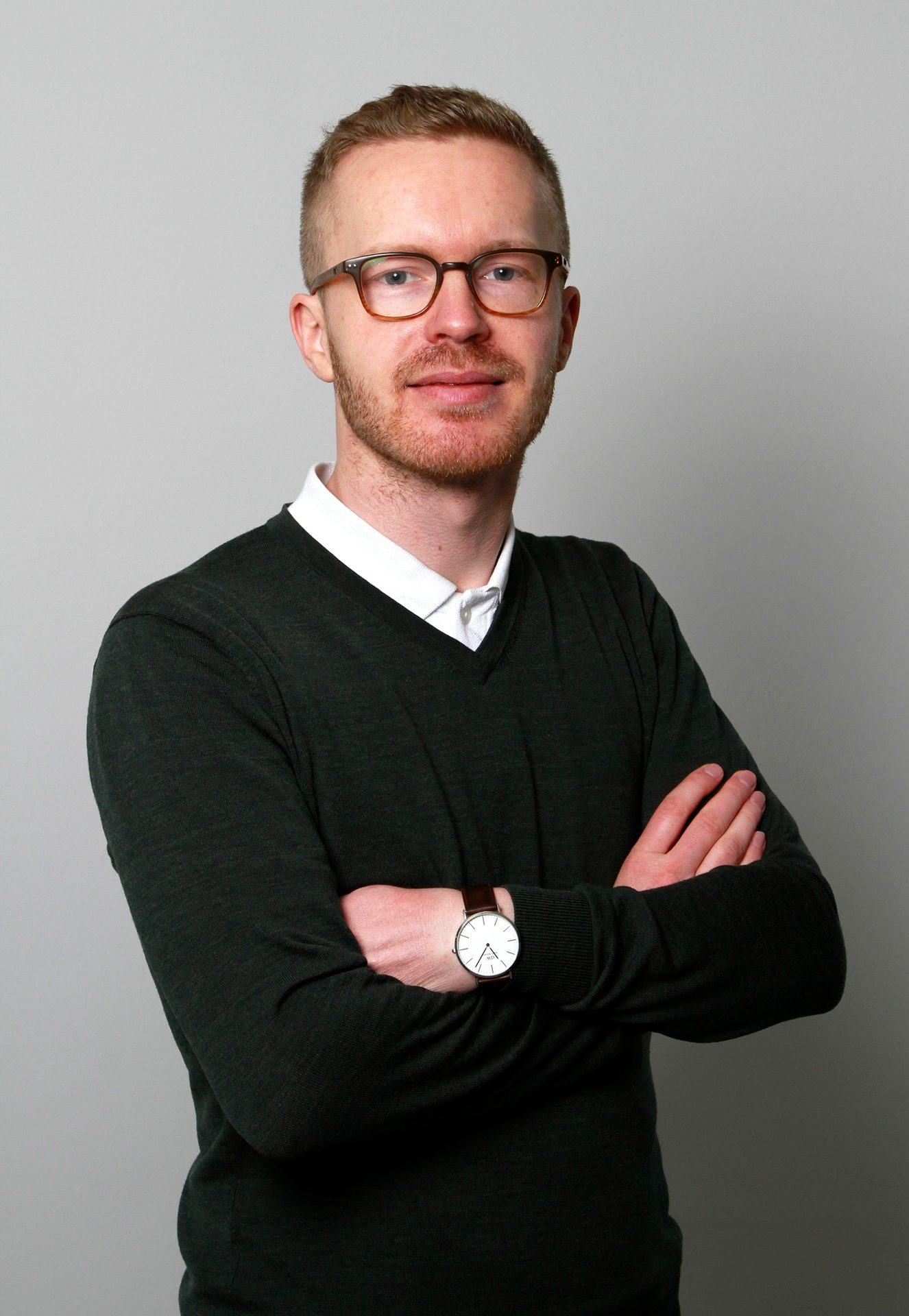 A man with glasses and a beard, wearing a green sweater and white shirt, stands with arms crossed.