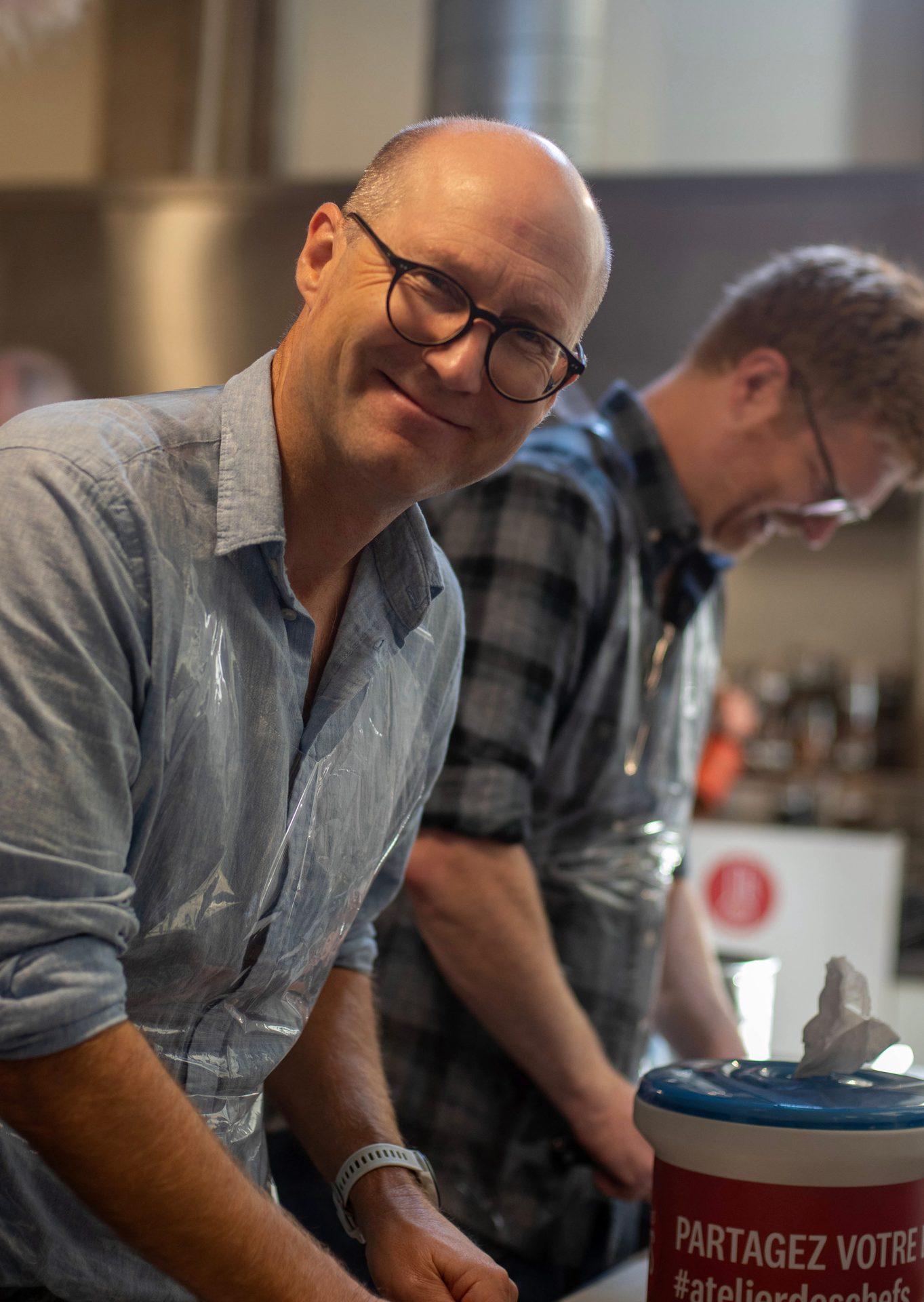 Smiling man in glasses and blue shirt with a plastic apron, looking at the camera.