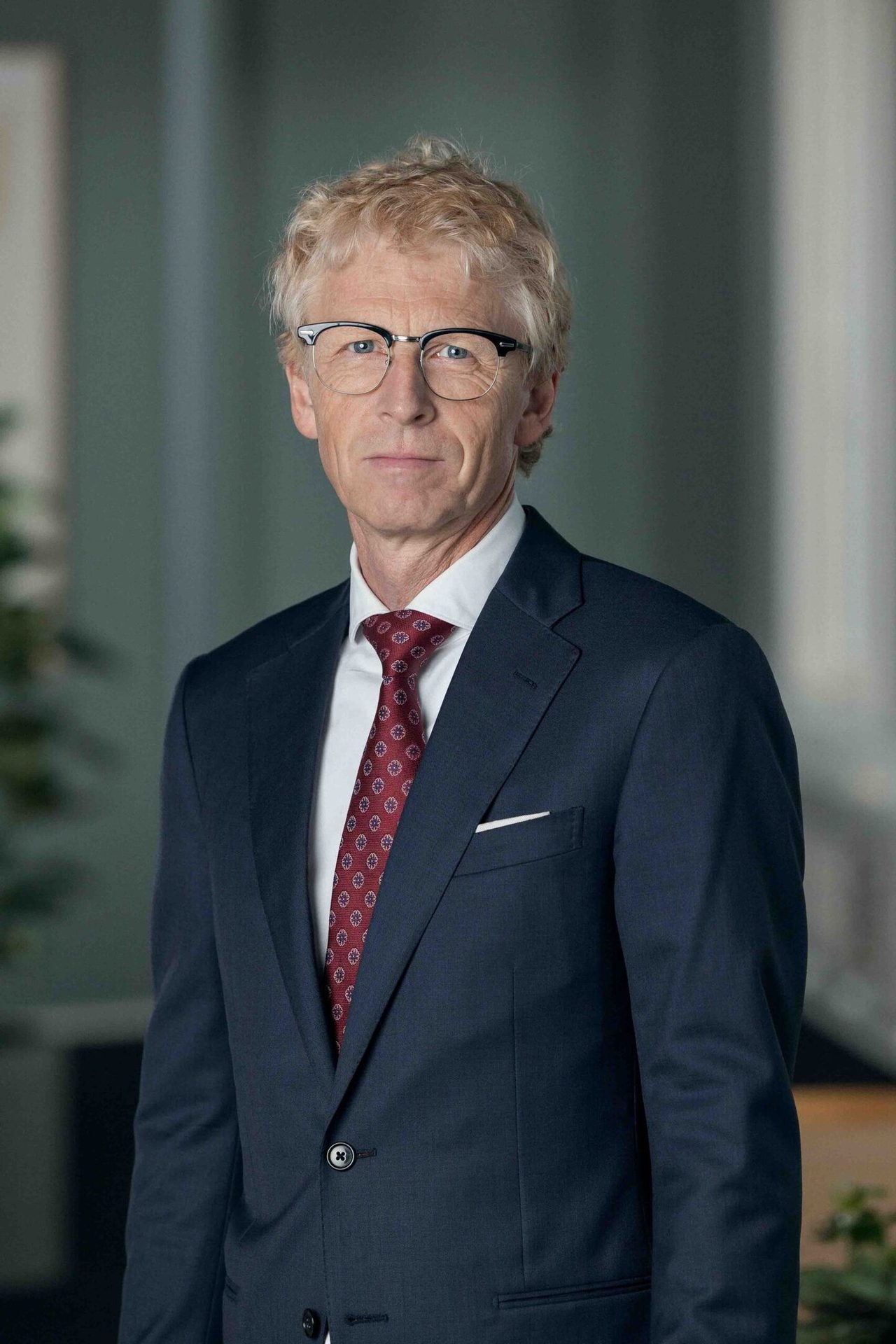 Professional portrait of a man with blonde hair and glasses, wearing a navy suit and red tie.