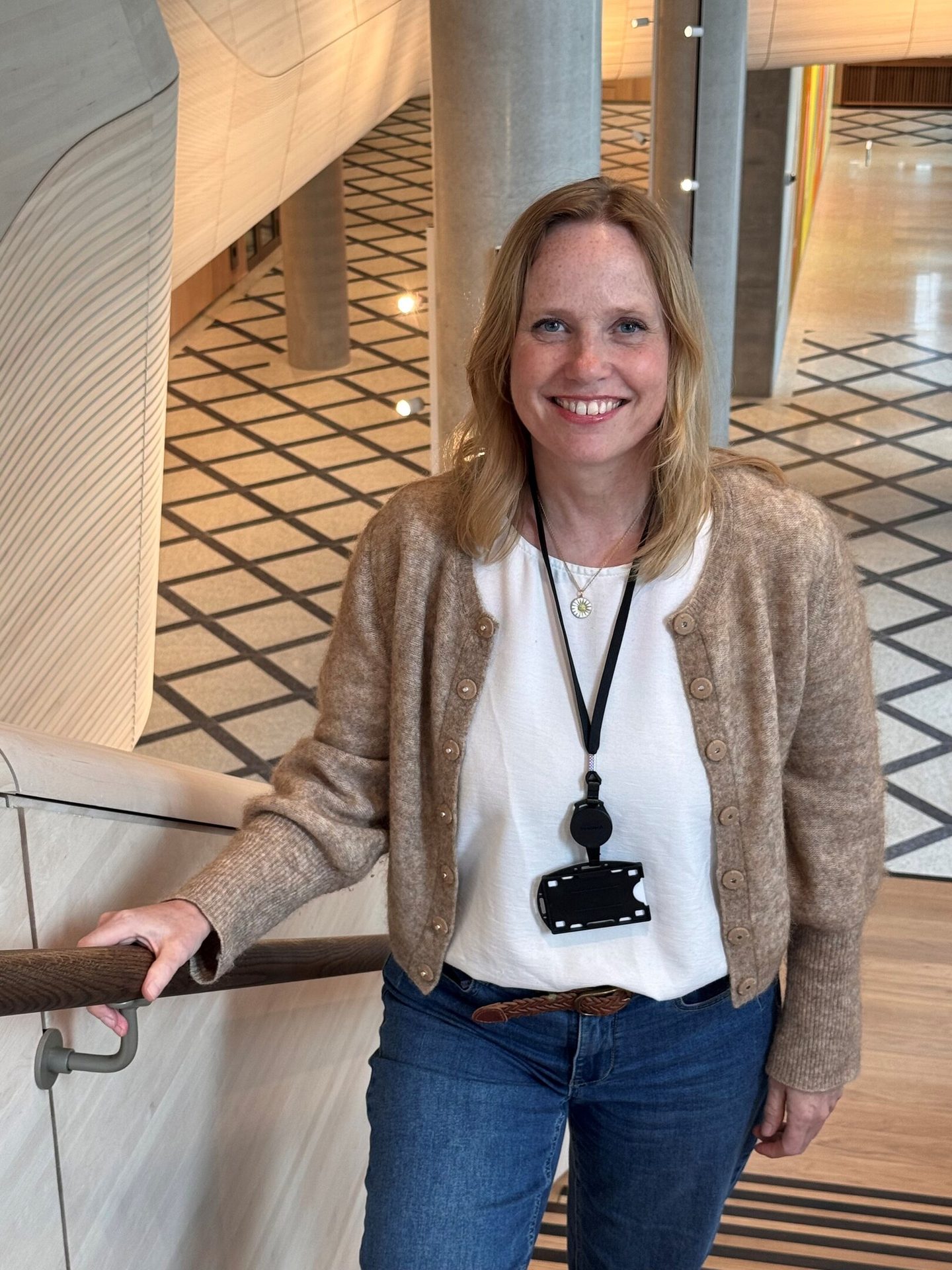 Smiling woman in white shirt and brown cardigan stands by railing indoors.