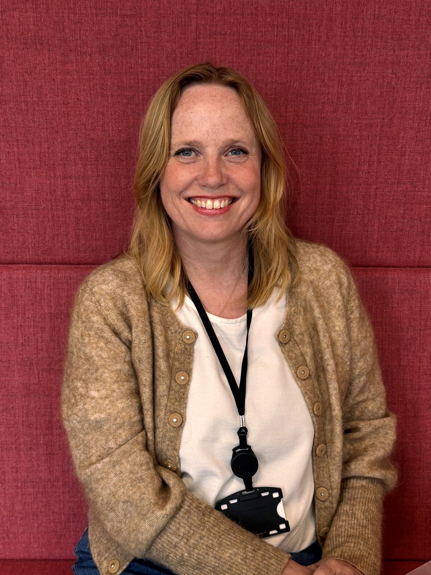 Smiling woman with blonde hair, wearing a beige cardigan and an ID badge, against a red textured background.