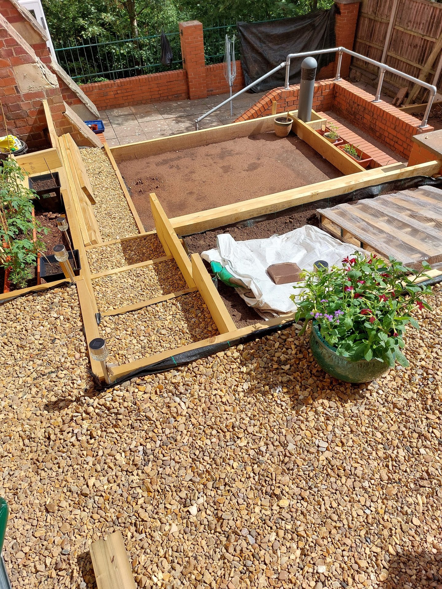 Garden under construction with raised wooden beds, gravel paths, brickwork, and potted plants.