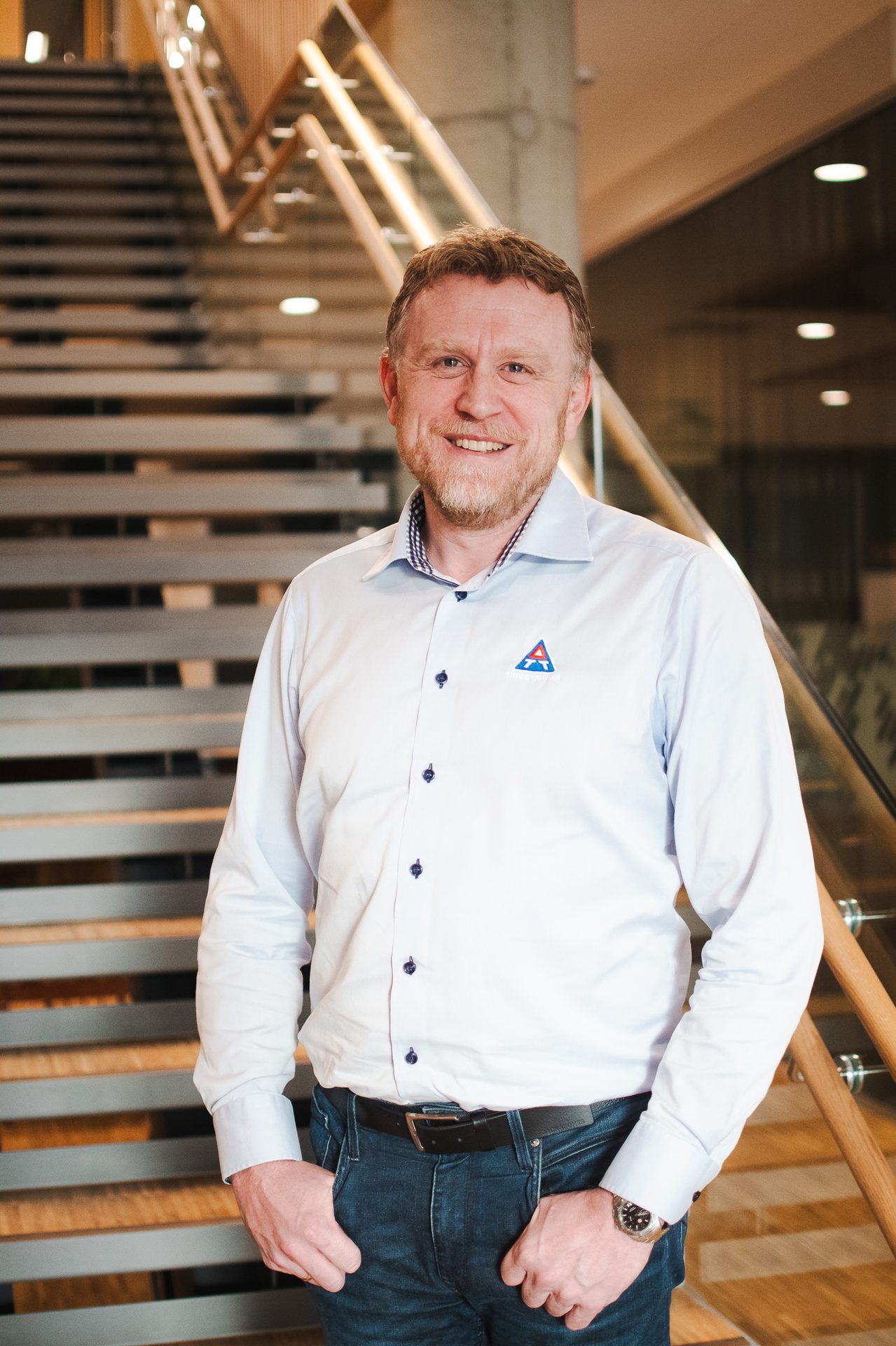 Smiling man with beard in light blue shirt, jeans, and watch, standing by a modern staircase.