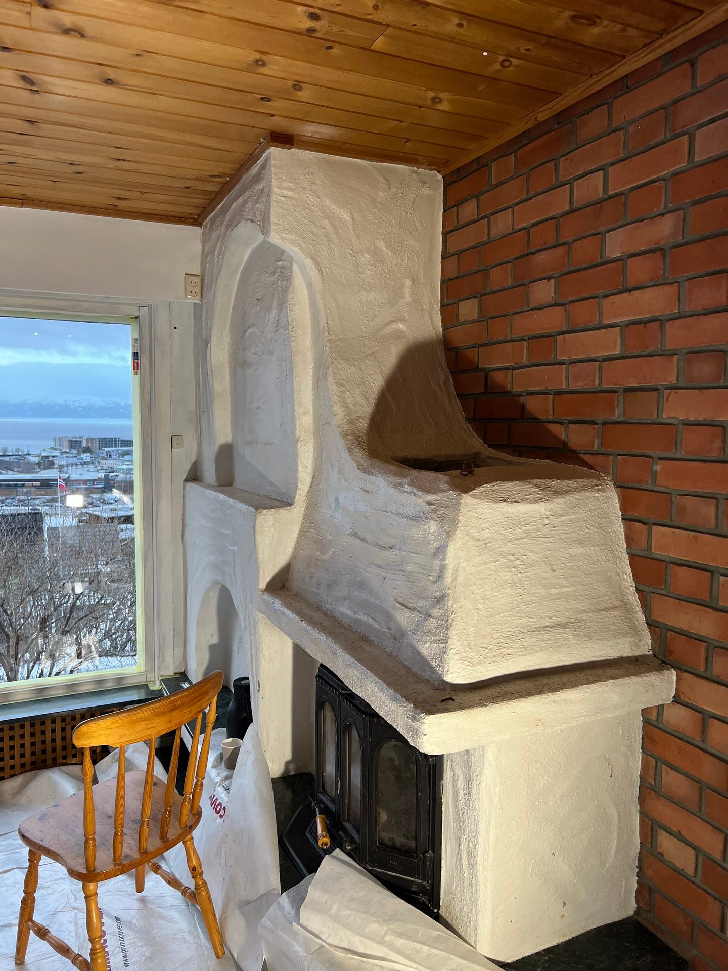 White fireplace, wood stove, brick wall, wood ceiling, chair, and window with a snowy outdoor view.