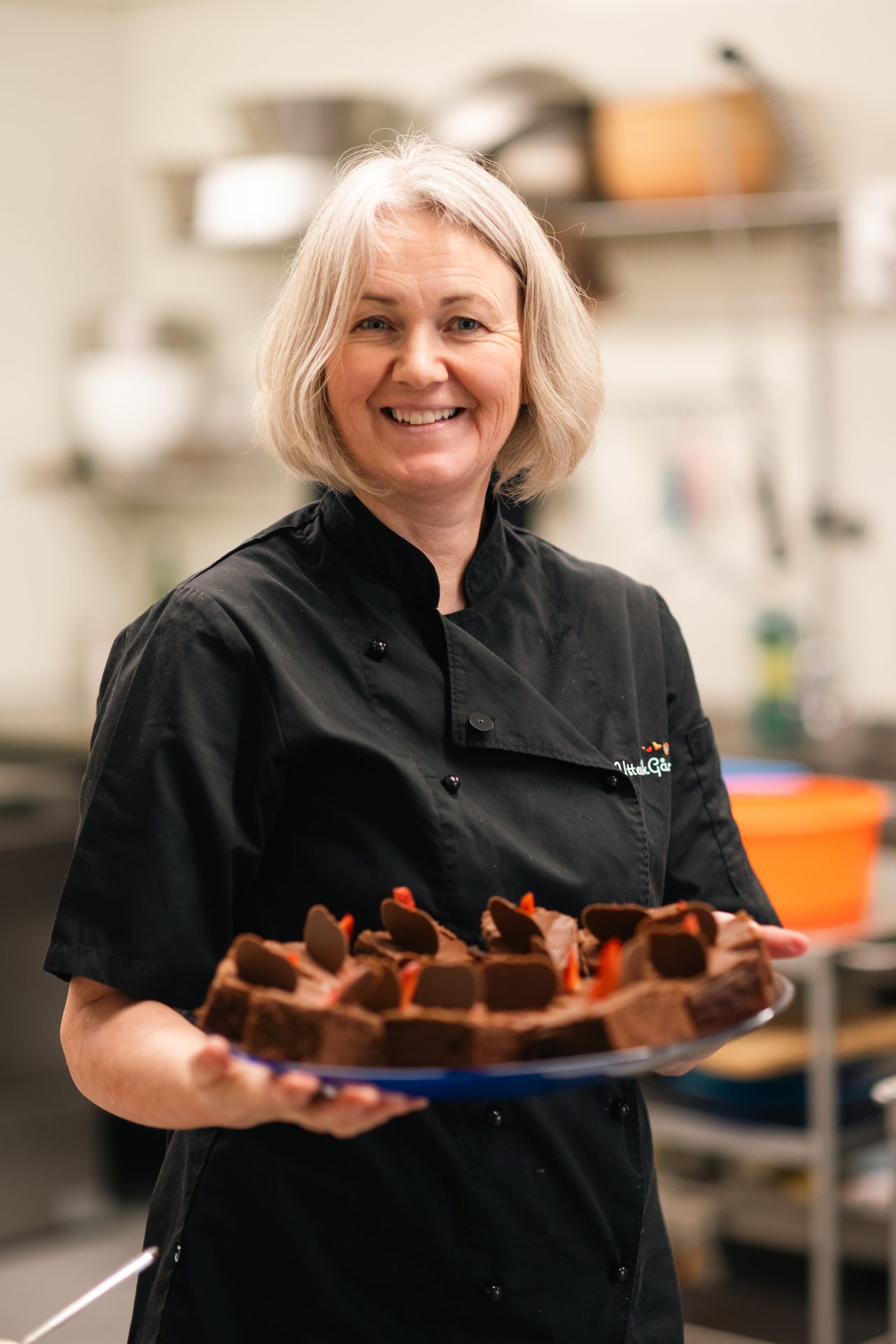 Smiling woman in chef's uniform holds a platter of chocolate cakes.