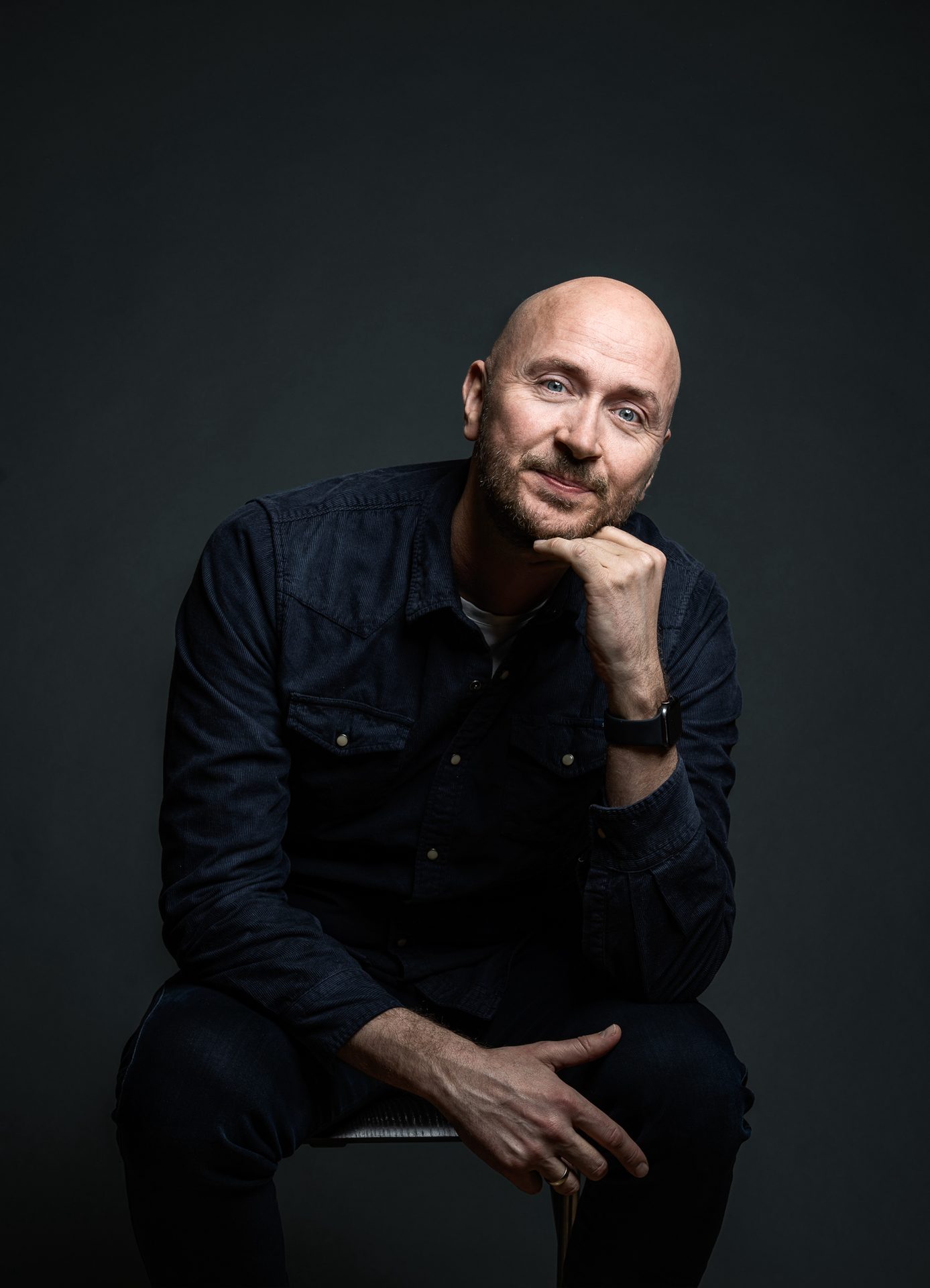 Bald, bearded man in dark shirt, sitting with chin in hand, looking forward.
