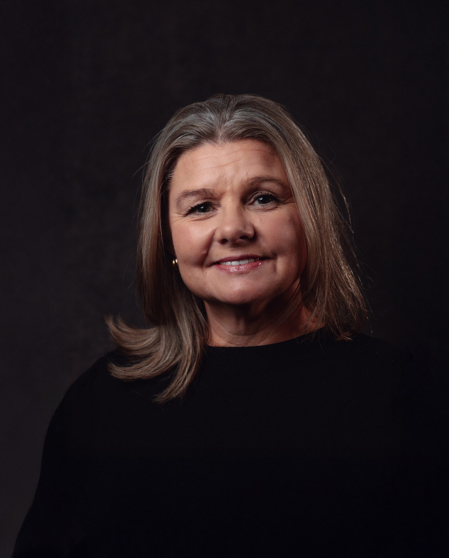 Smiling woman with gray and blonde hair in a black top, against a dark background.