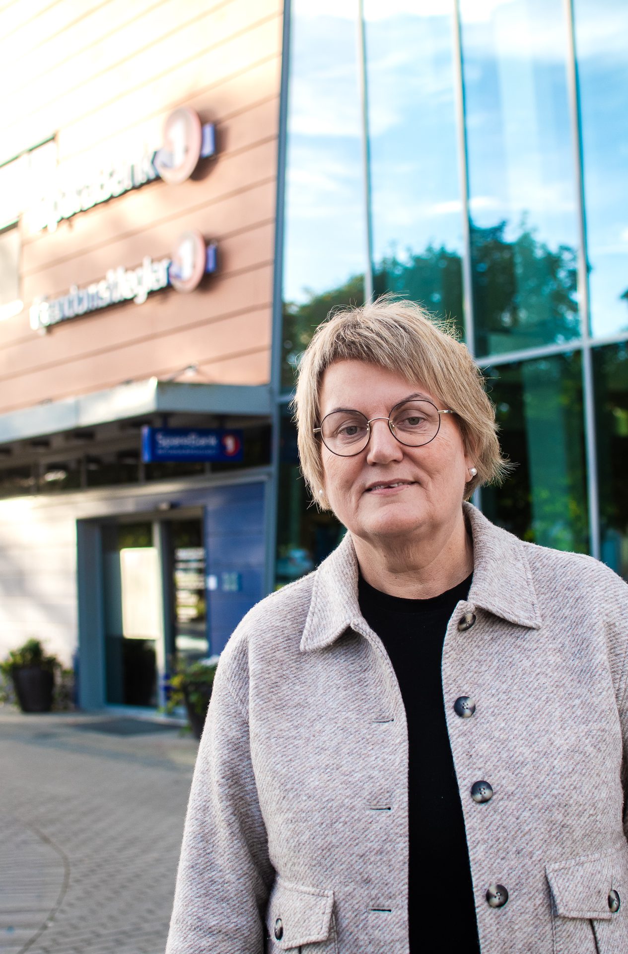 Older woman in glasses and light jacket stands outside a bank building with glass windows.
