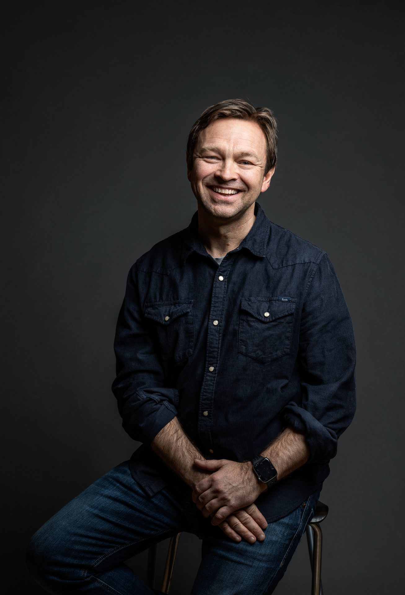 Smiling man in dark blue shirt and jeans, sitting.