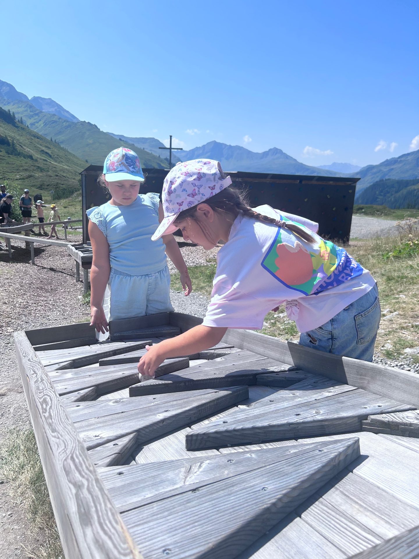 Two girls in caps playing with a wooden water maze outdoors in mountains under a clear sky.