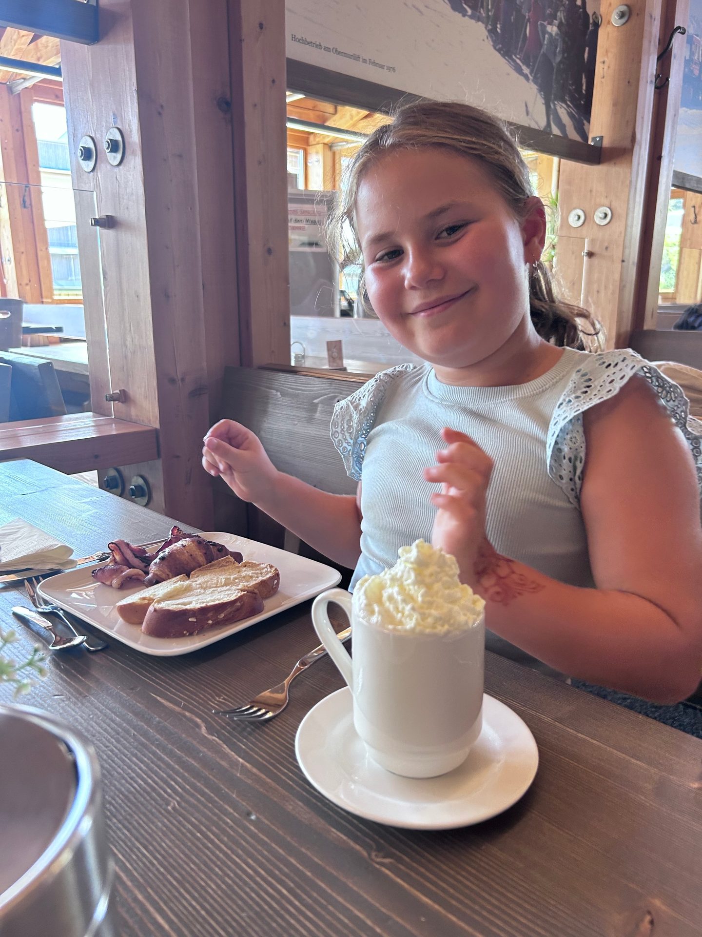 Young girl smiling at a table with breakfast and a drink with whipped cream in a rustic restaurant setting.