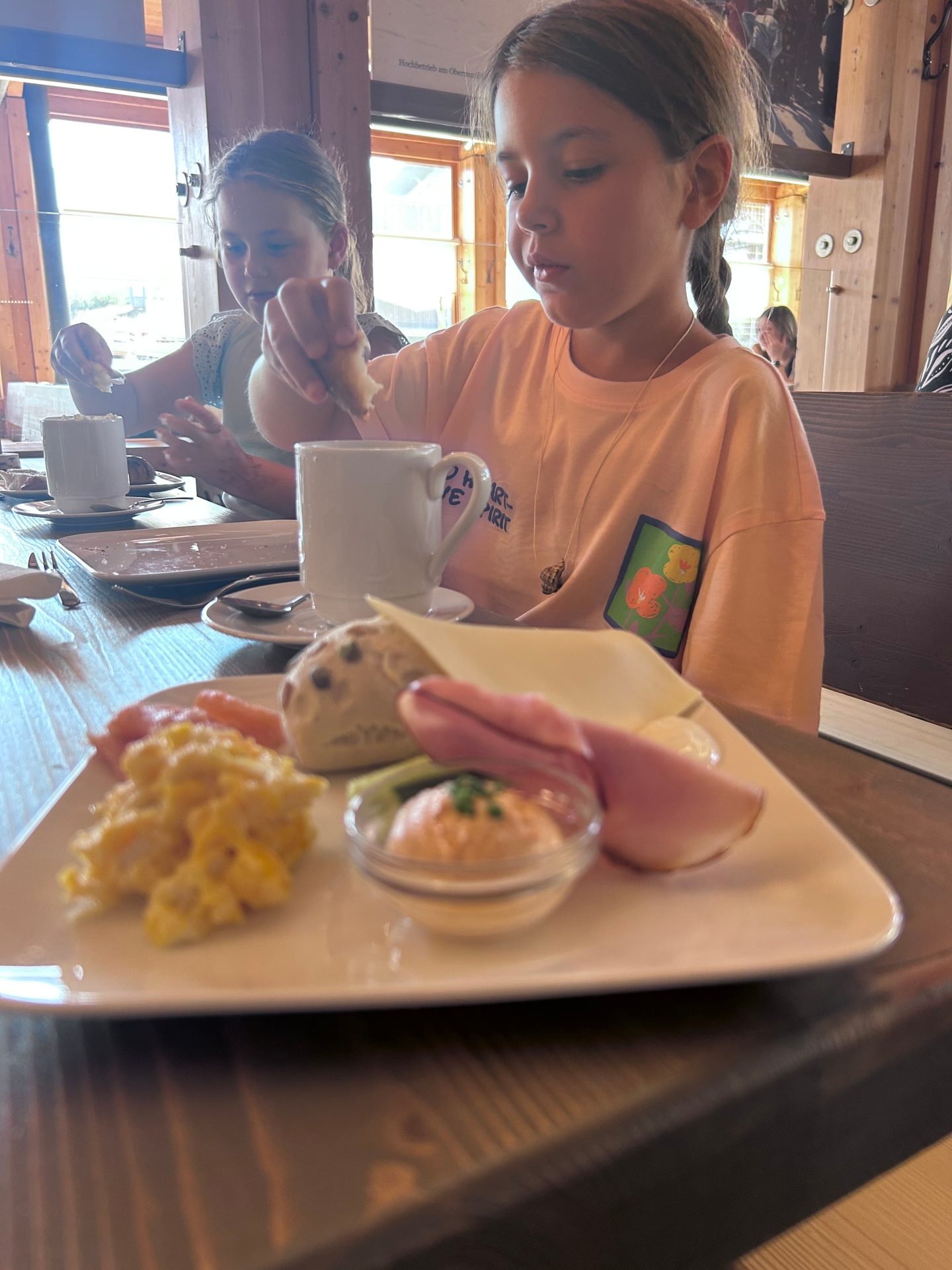 Two young girls enjoying breakfast at a table with a plate of eggs, ham, and bread.