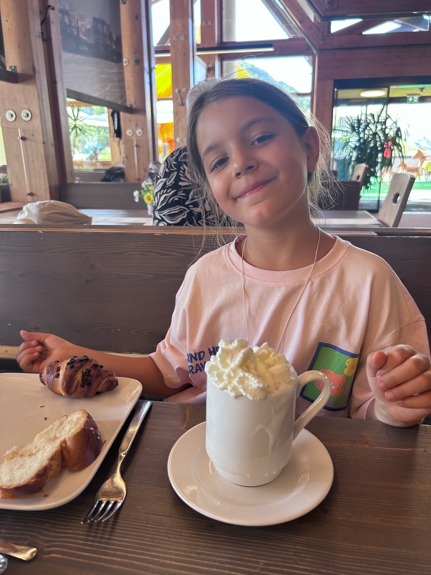 A young girl smiles at the camera while sitting at a table with a mug of whipped cream and pastries.