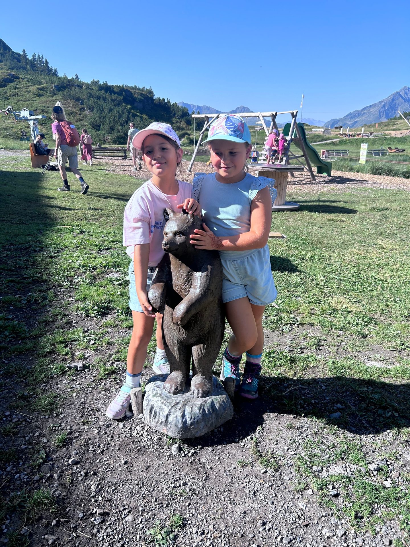 Two girls in hats pose with a carved wooden bear statue in a sunny mountain playground.