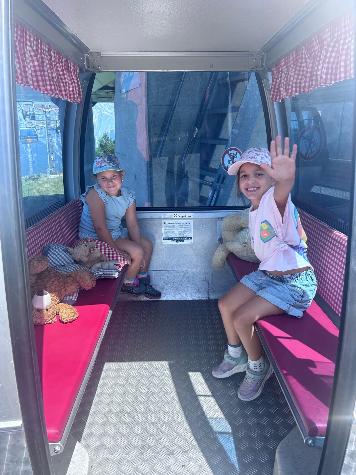 Two girls smile and wave from inside a cable car with teddy bears, enjoying the scenic ride.