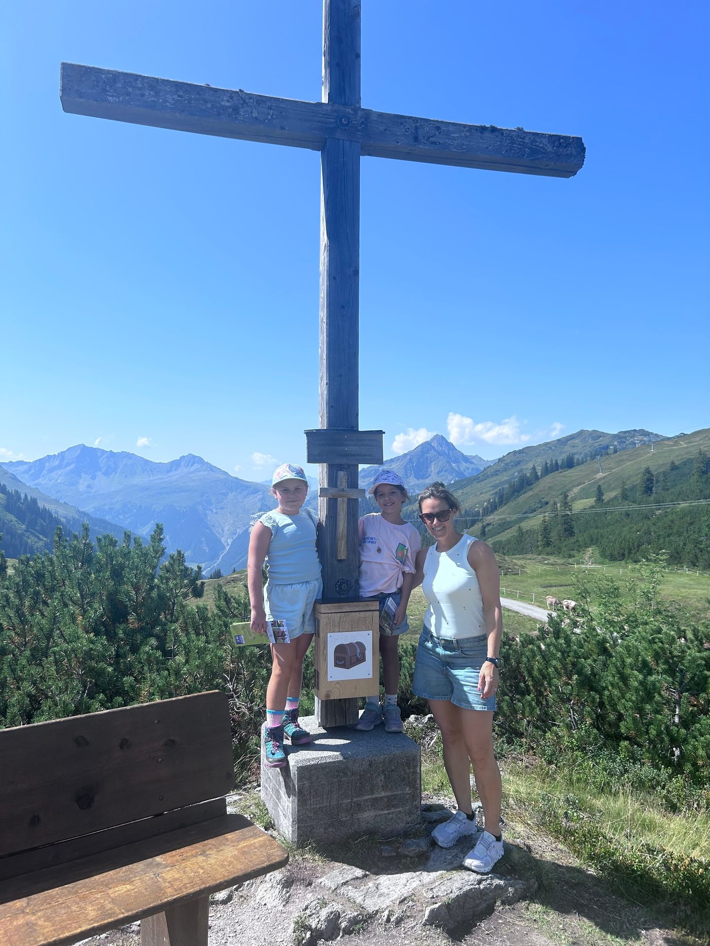 A woman and two girls pose at the base of a large wooden summit cross, with mountains in the background.