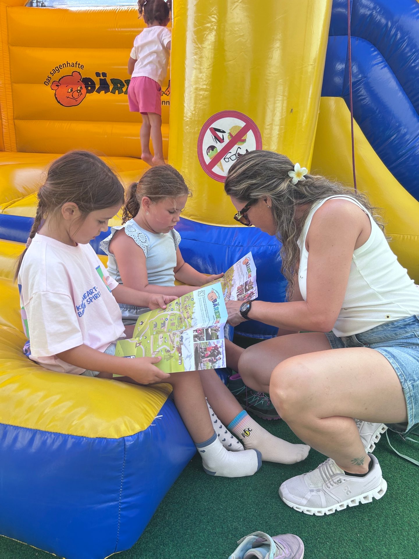 Two girls and a woman on a bouncy castle intently read a map or brochure together, with another child playing in the background.