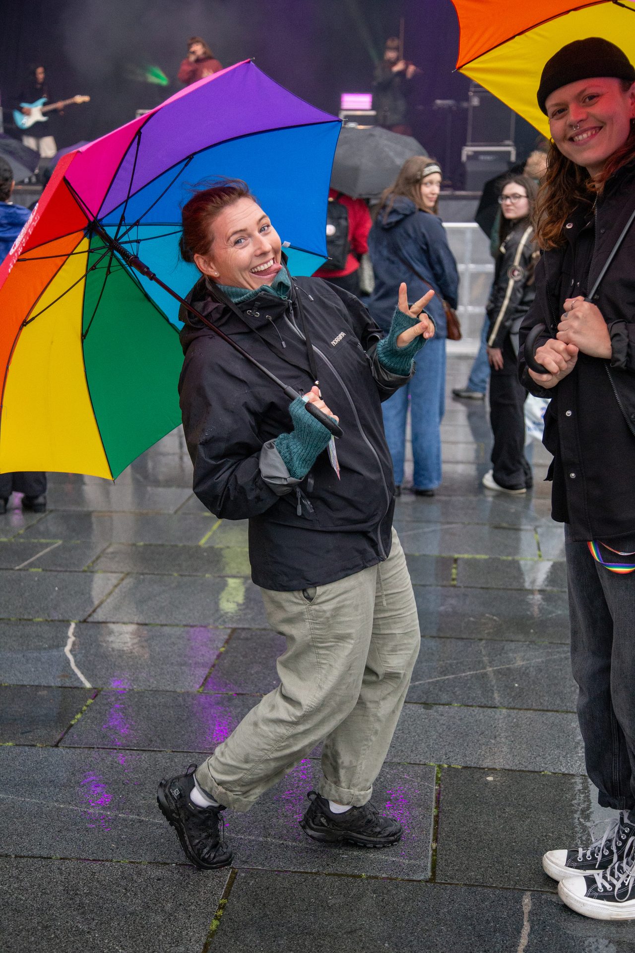 A woman with a rainbow umbrella makes a funny face, peace sign, and poses with another person.