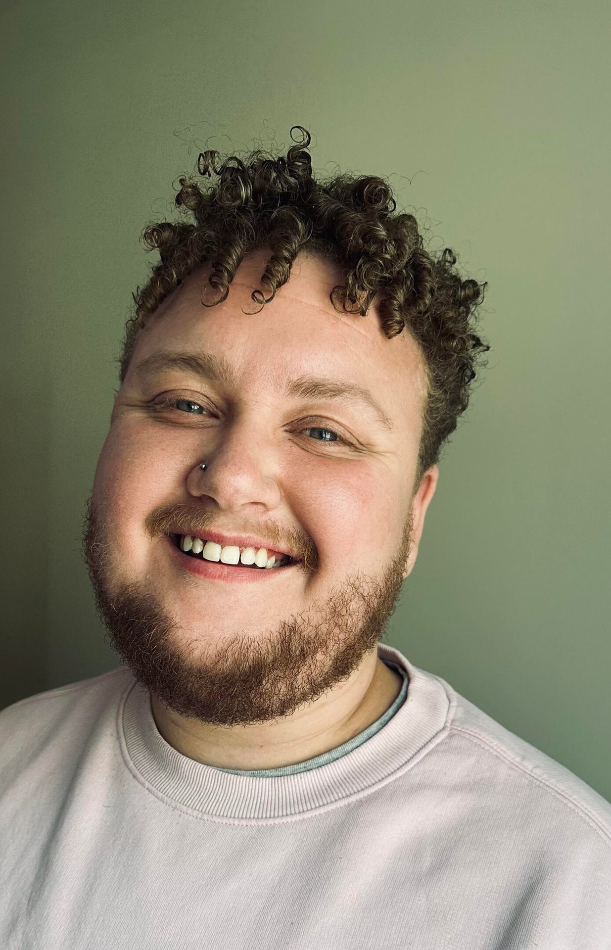 A smiling person with curly brown hair, a beard, and a nose piercing looks at the camera.