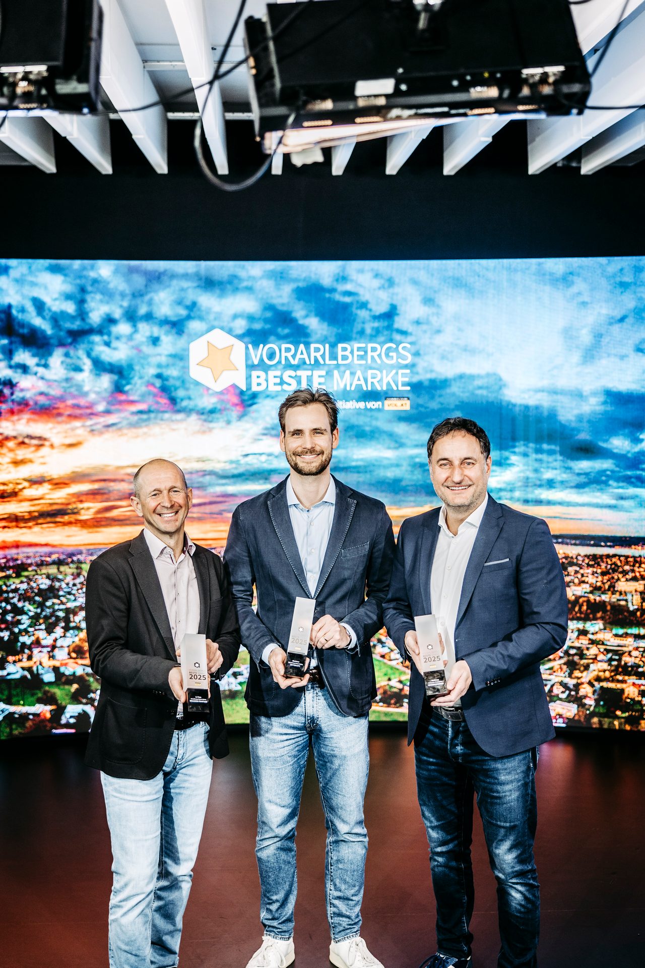 Three smiling men hold awards for "Vorarlbergs Beste Marke 2025" on a stage with a digital backdrop.