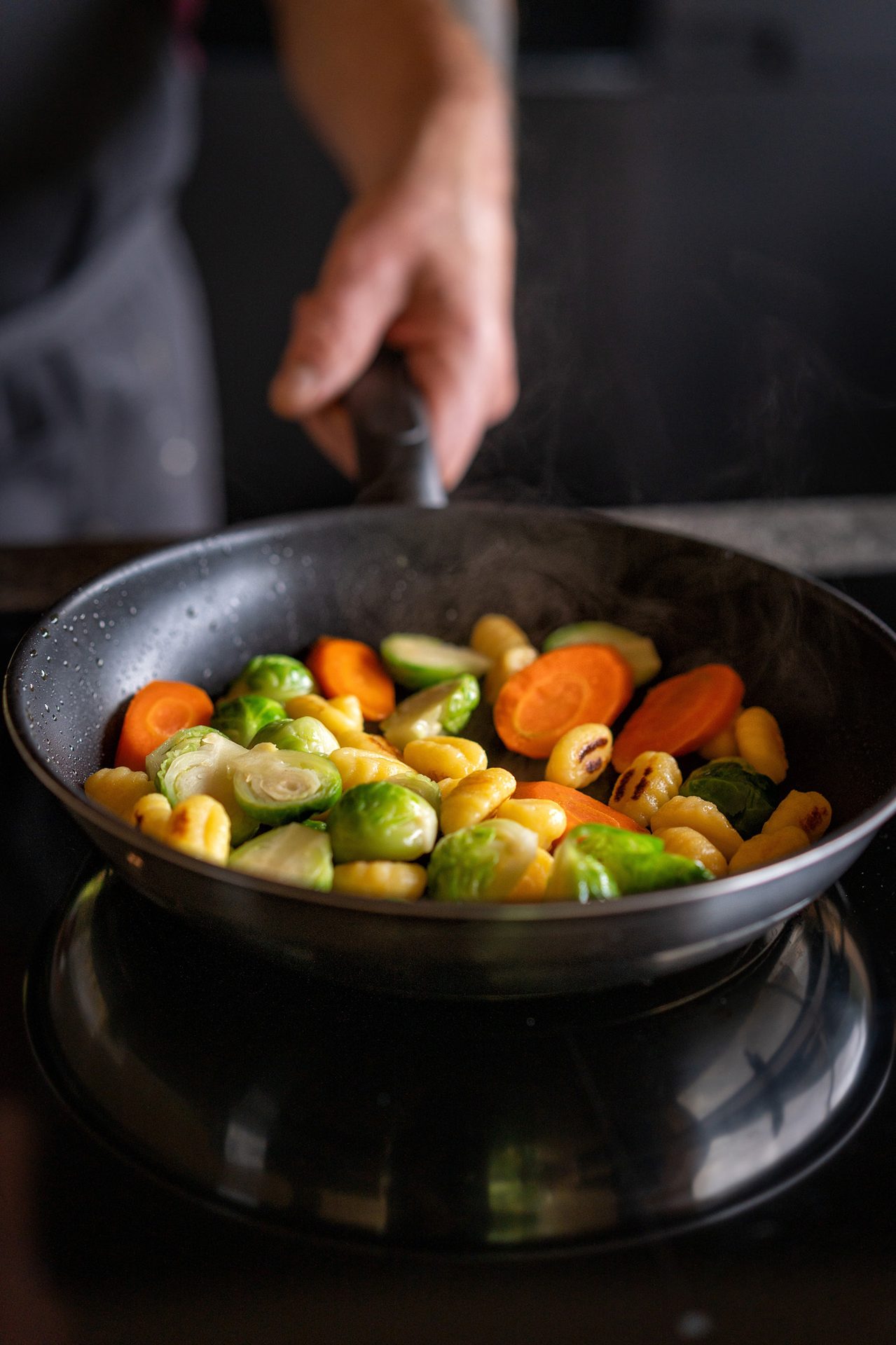 A hand holds a pan of gnocchi, Brussels sprouts, and carrots cooking on a black stovetop.