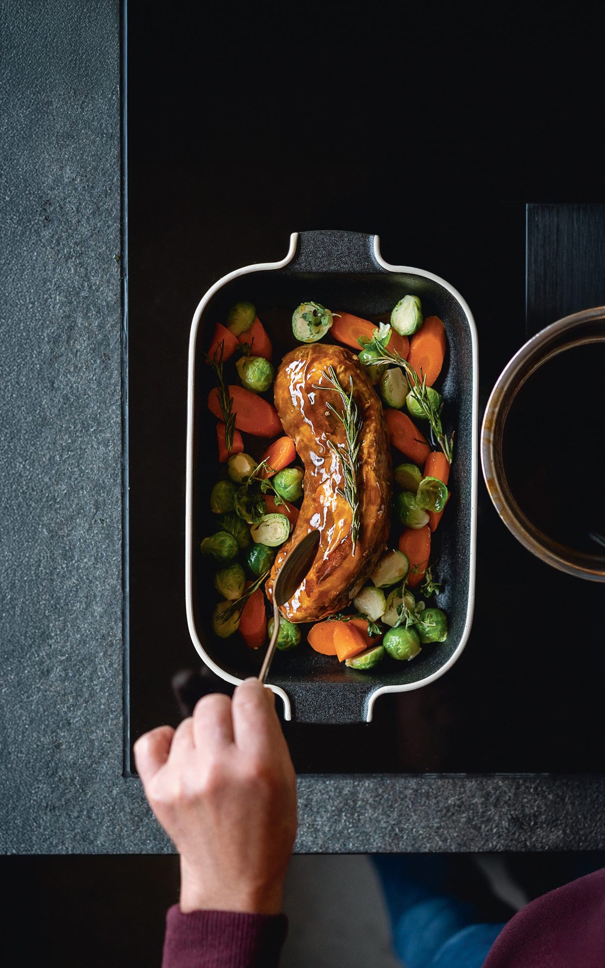 A hand serving glazed meat with roasted carrots and Brussels sprouts from a black roasting pan.
