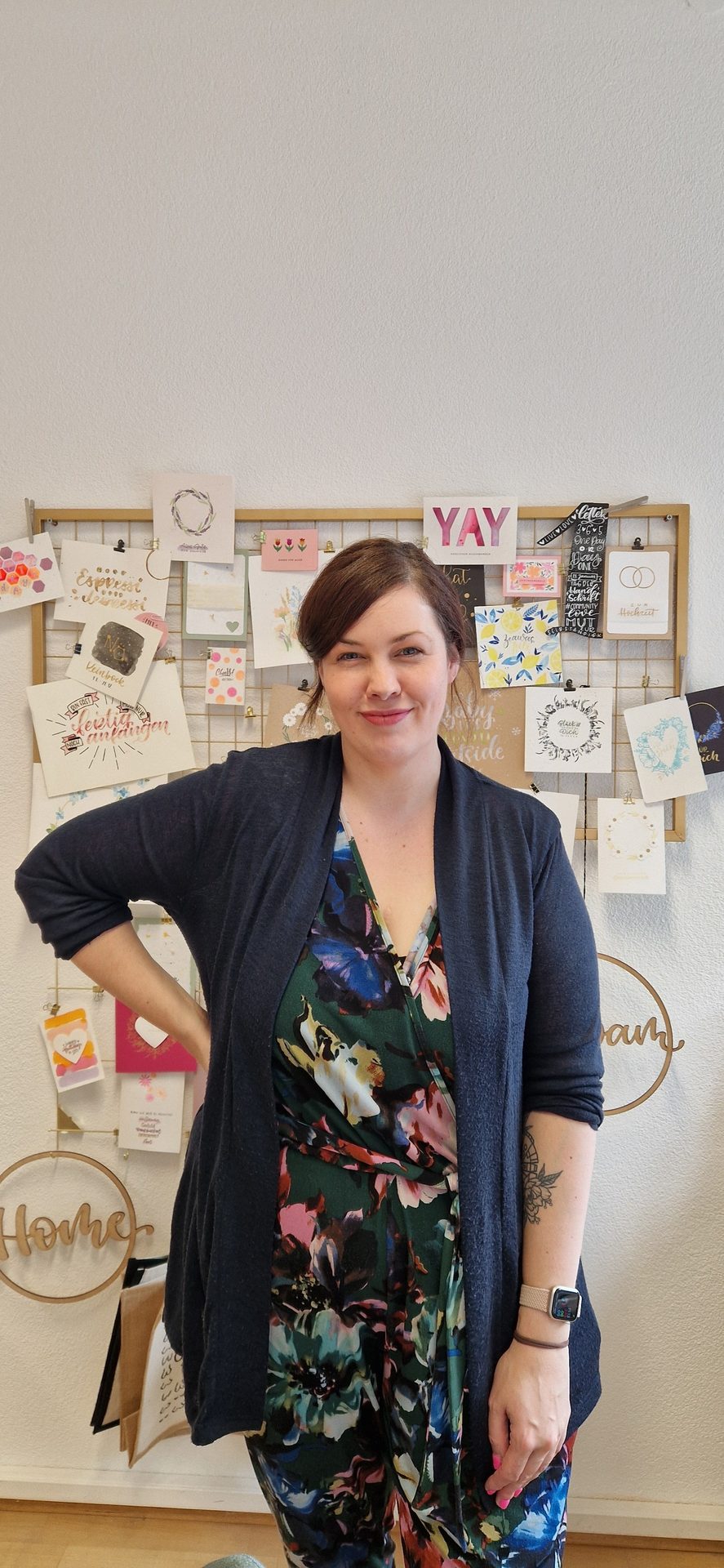 Smiling woman in floral dress and cardigan, standing in front of a grid board with cards.