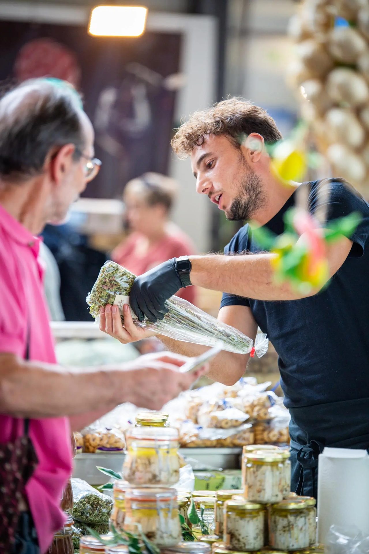 A vendor in gloves presents fresh herbs to a customer at an indoor market stall.